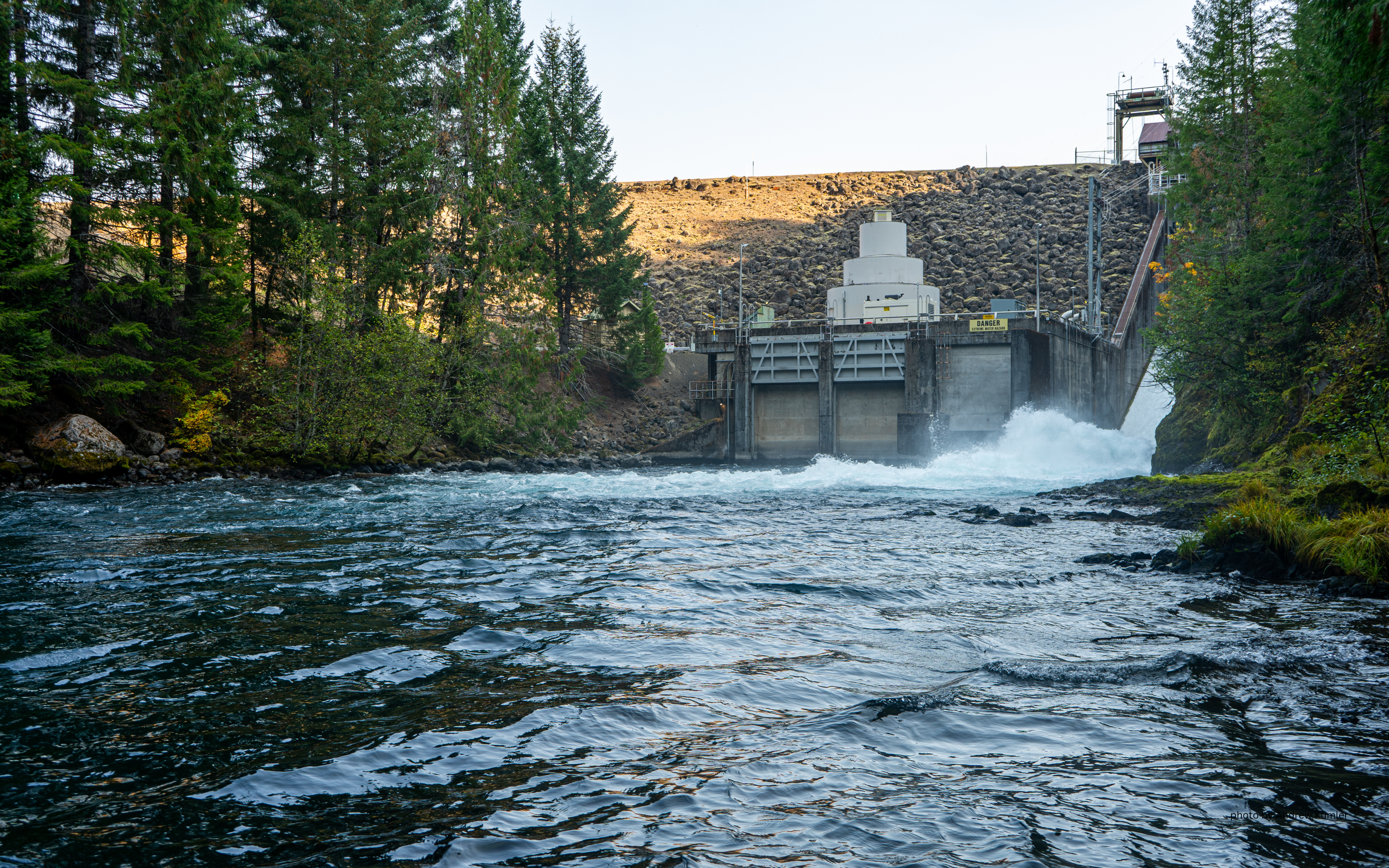 The Trail Bridge dam sits at the end of the frame. The dark blue water of the Mckenzie runs with green trees on the shore.