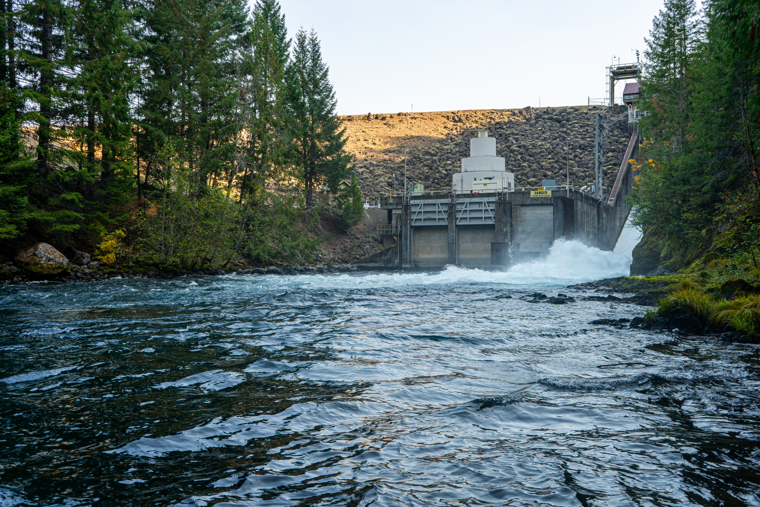 The Trail Bridge dam sits at the end of the frame. The dark blue water of the Mckenzie runs with green trees on the shore.